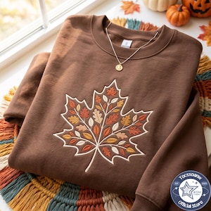 May include: A brown crewneck sweatshirt featuring a large embroidered maple leaf design on the front. The leaf is composed of smaller, multi-colored leaves in shades of orange, yellow, cream, and rust. A thin gold chain necklace rests on the collar.