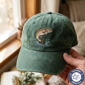 Könnte beinhalten: Eine waldgrüne Baseballkappe mit einer Stickerei einer springenden Forelle in Rosa-, Beige- und Brauntönen, mit hellblauen Wasserspritzern unter dem Fisch. Die Kappe hat eine strukturierte Krone und einen gebogenen Schirm.