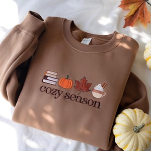 May include: A brown sweatshirt with embroidered fall-themed designs, including books, a pumpkin, a maple leaf, and a coffee cup. The words "cozy season" are embroidered below the designs. The sweatshirt is surrounded by fall leaves and small pumpkins.