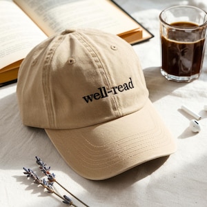May include: A tan baseball cap with the embroidered text "well-read" in black. The cap is on a white surface next to a glass of coffee, an open book, and a pair of white earbuds. The cap has a curved brim.