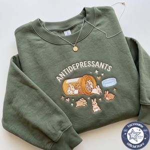May include: An olive green crewneck sweatshirt featuring an embroidered graphic of an orange pill bottle labelled ANTIDEPRESSANTS, with several small brown and white bunnies spilling out of it, surrounded by small embroidered hearts and stars.