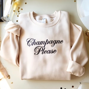 May include: A light beige sweatshirt with the embroidered phrase "Champagne Please" in elegant black script. The crew neck sweatshirt has long sleeves and is folded neatly. The image has a celebratory theme.