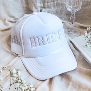 May include: A white trucker hat with the word "BRIDE" embroidered in a light pink thread. The hat features a mesh back and a curved brim. A clear wine glass and a small book are in the background.