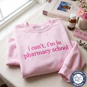 May include: A light pink crewneck sweatshirt featuring the embroidered text "i can't, i'm in pharmacy school" in a dark pink thread. The garment has long sleeves with ribbed cuffs and a ribbed neckline, showcasing a simple and clean design.