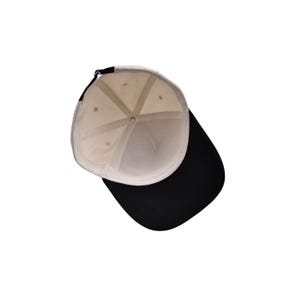 May include: A baseball cap with a black brim and a cream-colored crown. The interior of the cap is cream-colored with a black adjustable strap. The cap is viewed from the inside.