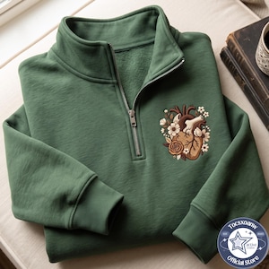 Puede incluir: Una sudadera verde bosque con cremallera de un cuarto, cuello alto y puños acanalados. Un diseño bordado en el pecho izquierdo muestra un corazón anatómico en tonos marrones, rodeado de pequeños detalles florales en blanco y crema.