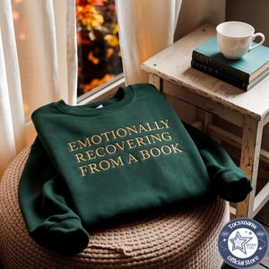 Puede incluir: Una sudadera de cuello redondo en color verde bosque oscuro con el texto bordado "EMOTIONALLY RECOVERING FROM A BOOK" en hilo de color dorado. La prenda tiene mangas largas, un cuello acanalado y una construcci&oacute;n de tela suave y lisa.