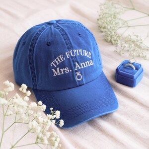 May include: A blue baseball cap embroidered with the text "THE FUTURE Mrs. Anna" and a ring symbol. A blue velvet ring box holds a diamond ring. White flowers are arranged around the cap and box.