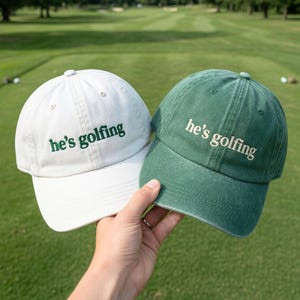 May include: Two baseball caps, one white and one green, are held up against a golf course background. Both caps have the phrase "he's golfing" embroidered in green. The white cap has a white brim, and the green cap has a green brim.