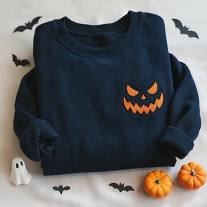 Puede incluir: Sudadera azul marino con un bordado naranja de una cara de calabaza de Halloween. La prenda doblada está decorada con murciélagos de papel y pequeñas calabazas, creando una escena festiva. Las mangas están metidas, presentando una estética otoñal.