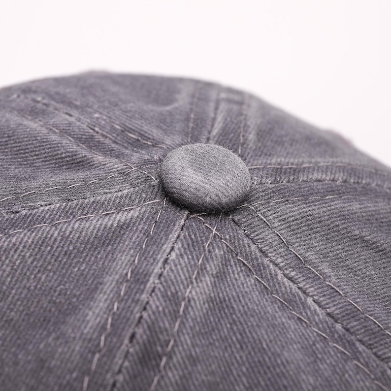 May include: Close-up of a gray newsboy cap with a button on top. The cap is made of a textured fabric with visible stitching. The button is a matching gray color.