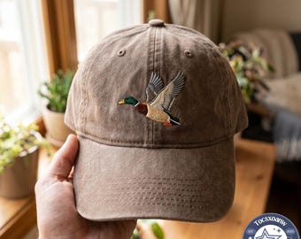 Puede incluir: Una gorra de béisbol marrón con una textura desgastada y lavada. El panel frontal presenta un diseño bordado de un pato real en vuelo, mostrando un plumaje detallado en verde, marrón, blanco y gris con pico y patas naranjas.