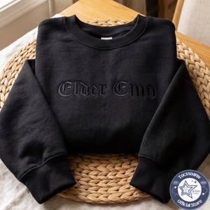 May include: A black crewneck sweatshirt featuring the text Elder Emo embroidered in a matching black thread across the chest. The garment has long sleeves with ribbed cuffs and a ribbed neckline, showcasing a clean and uniform monochromatic design.