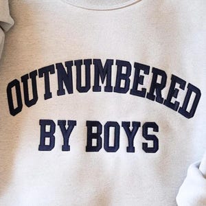 May include: A light gray sweatshirt with the navy blue text "OUTNUMBERED BY BOYS" in a collegiate font. The sweatshirt has a crew neck and long sleeves. The text is embroidered on the front.