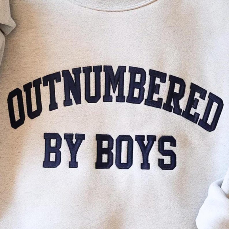 May include: A light gray sweatshirt with the navy blue text "OUTNUMBERED BY BOYS" in a collegiate font. The sweatshirt has a crew neck and long sleeves. The text is embroidered on the front.