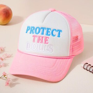 May include: A pink and white trucker hat with the embroidered text "PROTECT THE DOLLS" in blue, pink, and white. The hat has a pink peak and mesh back. The hat has a curved peak and a button on top.