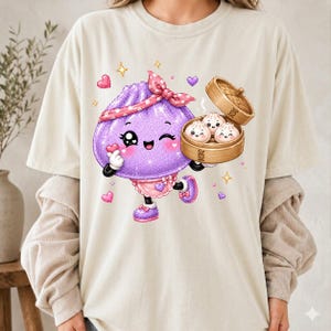 May include: A cream-colored t-shirt featuring a graphic of a purple, glittery, anthropomorphic dumpling character wearing a pink polka-dot headband and apron, holding a wooden steamer basket containing three smaller smiling dumpling characters.