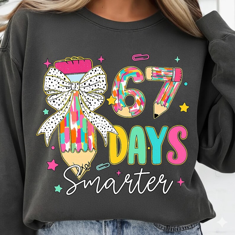 May include: Dark grey sweatshirt featuring a colourful pencil graphic and the text "67 Days Smarter." The pencil design includes a polka dot bow and a rainbow of colours. The sweatshirt is a comfortable, casual style.