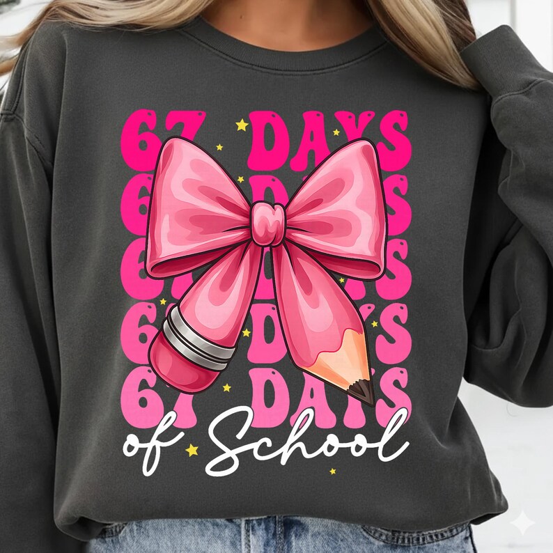 May include: Dark grey sweatshirt featuring a pink bow and pencil design. The text reads "67 Days of School" in pink and white. The design includes stars and a retro font, suitable for back-to-school or teacher gifts.