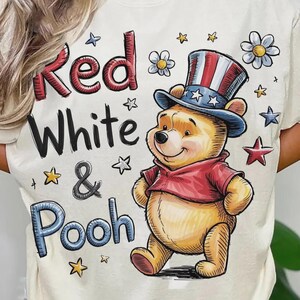 Könnte beinhalten: Cremefarbenes T-Shirt mit einem Cartoon-Bären, der einen patriotischen Zylinder und ein rotes Hemd trägt. Der Text "Red White & Pooh" ist über und um den Bären gedruckt, mit Sternen und Blumen verstreut.