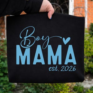 May include: Black tote bag with the words "Boy MAMA" in light blue script and block letters, with a small heart symbol. The text "est. 2026" is below the word "MAMA".