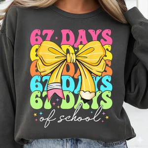 May include: Dark grey sweatshirt with a colourful graphic. The graphic features the text "67 DAYS of school" in a stacked design with a yellow bow and a pencil. The text is in pink, yellow, orange, blue, and green.