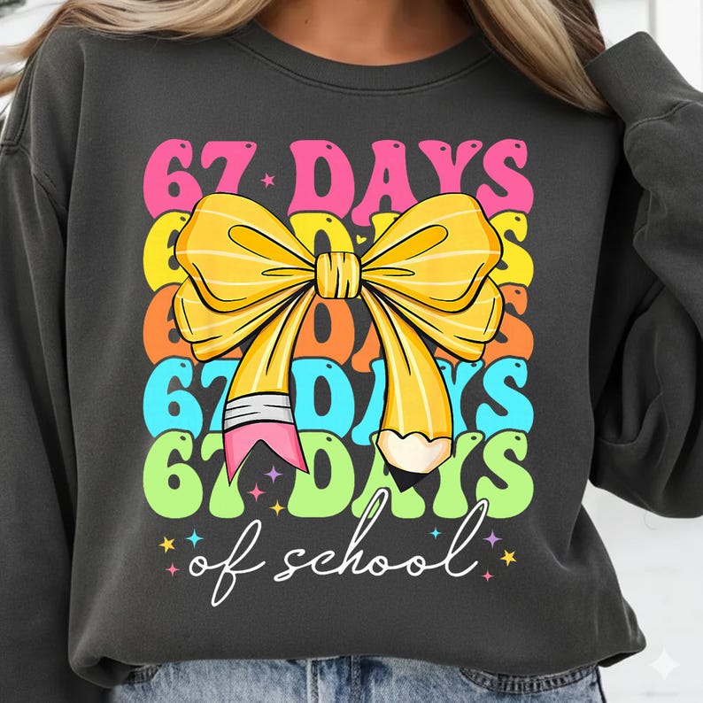 May include: Dark grey sweatshirt with a colourful graphic. The graphic features the text "67 DAYS of school" in a stacked design with a yellow bow and a pencil. The text is in pink, yellow, orange, blue, and green.