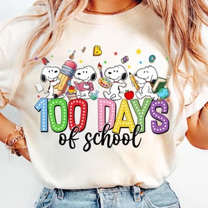 以下が含まれることがあります： クリーム色のTシャツ。スヌーピーのキャラクターが100日間の学校を祝っています。デザインには、鉛筆、本、絵の具を持ったスヌーピーと、カラフルな文字で「100 Days of school」というテキストが含まれています。このシャツは楽しく、教育的なテーマです。