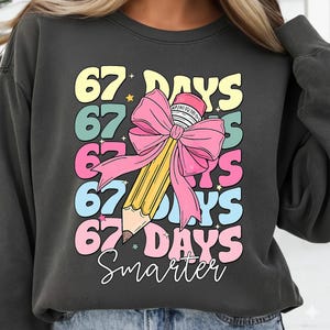May include: Dark grey sweatshirt with a colourful graphic. The design features the text "67 DAYS" in various colours, a large yellow pencil with a pink bow, and the word "Smarter" in a script font. A star is also included.