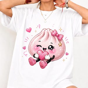 May include: A white short-sleeve t-shirt featuring a graphic of a pink, glittery, cartoon-style dumpling character with a winking face, wearing a pink bow, and holding a pink heart, surrounded by small pink hearts and gold star accents.
