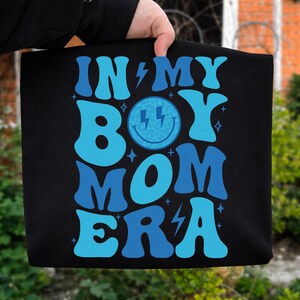 May include: Black tote bag with the text "IN MY BOY MOM ERA" in shades of blue. The design includes a smiley face with lightning bolts and star accents. The bag is held by a person.
