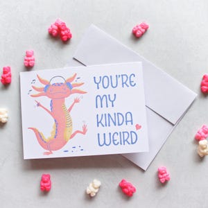 May include: A greeting card with a dancing axolotl wearing headphones, the text reads "YOU'RE MY KINDA WEIRD". The card is accompanied by a white envelope and scattered pink and white gummy bears.