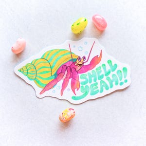 May include: A colorful sticker featuring a cartoon hermit crab with a pink body and a yellow and green striped shell. The sticker has the words "SHELL YEAH!!" in green. Several jelly beans surround the sticker.