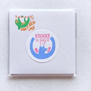 May include: A square pack of sticky notes with a green sloth graphic and the words "Don't Rush Me." A circular sticker features a blue bear with pink flowers and the text "Stickies 50 Sheets 3 x 3".