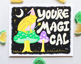 May include: A black background with white glitter and stars. A yellow frog wearing a purple hat sits on a green mushroom with white spots. A purple mushroom with white spots is behind the frog. The text "You're Magical" is written in white with a yellow outline.