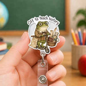 Peut inclure: Un enrouleur de badge circulaire illustré d'une grenouille verte portant des lunettes et un costume marron, sur une trottinette. La grenouille porte un panier avec des livres et un globe. Le texte au-dessus indique, Off to teach history.