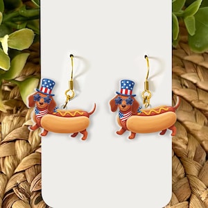 May include: A pair of novelty earrings featuring dachshund dogs dressed as hot dogs. The dogs wear sunglasses, top hats, and patriotic bandanas. The earrings have gold-colored hooks and are set against a white background.