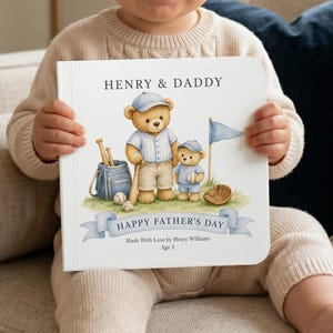 Op de afbeelding: Een gepersonaliseerd kinderboek met een honkbalthema. De omslag toont illustraties van teddyberen, honkbaluitrusting en de tekst "HENRY & DADDY" en "HAPPY FATHER'S DAY". Het boek wordt door een kind vastgehouden.