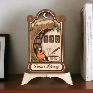 May include: Wooden library-themed decor with a cutout design. Features a girl reading, a tree, a bookshelf, and the text "Lucia's Library" and "Book Read This Year". The counter shows "120".