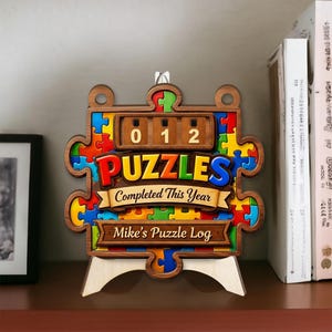 以下が含まれることがあります： カラフルな文字で「PUZZLES」と書かれた、木製のパズル型のディスプレイ。 「012」と表示されたカウンターと、「Completed This Year」と「Mike's Puzzle Log」のテキストがあります。 ディスプレイは木製のスタンドにあります。