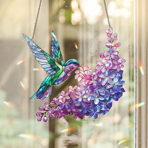 Puede incluir: Un colibrí de vidriera con alas iridiscentes y un pico largo, alimentándose de un racimo de flores moradas y rosas. El colibrí está suspendido por una cadena plateada, creando una decoración de ventana.