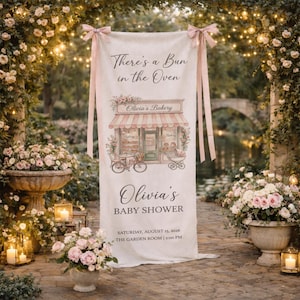 May include: A vertical fabric banner with pink ribbon ties at the top, featuring the text "There's a Bun in the Oven", "Olivia's Bakery", "Olivia's BABY SHOWER", and event details "SATURDAY, AUGUST 15, 2026" and "THE GARDEN ROOM | 2:00 PM".