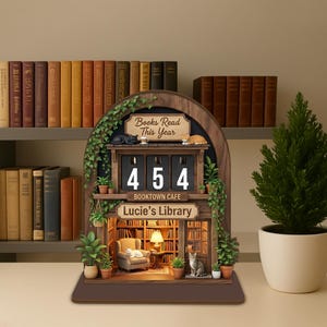 May include: A wooden decorative piece with a library theme. It features a counter displaying "454" books read, with the text "Books Read This Year" and "Lucie's Library." The piece includes miniature plants, cats, and a cozy reading nook.