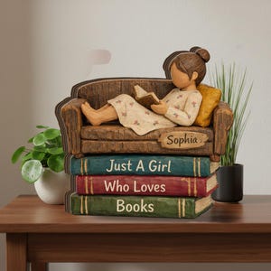 Może przedstawiać: Drewniana figurka dziewczyny czytającej na kanapie, na stosie książek. Książki głoszą "Just A Girl Who Loves Books". Kanapa ma żółtą poduszkę. Figurka jest spersonalizowana imieniem "Sophia".