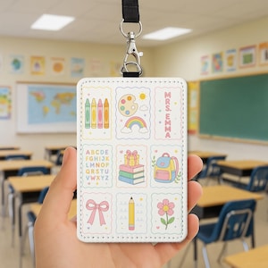 May include: A white rectangular ID badge holder with a black lanyard featuring a grid of colourful illustrations including crayons, a paint palette, a rainbow, the text MRS. EMMA, an alphabet, a stack of books with a gift, a backpack, a bow, a pencil, and a flower.