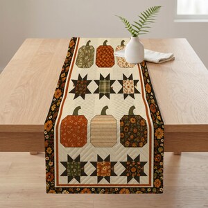 May include: A rectangular quilted table runner featuring a cream-colored background with a repeating pattern of pumpkins and star shapes in shades of orange, brown, green, and tan. The runner has a dark border with a floral print.
