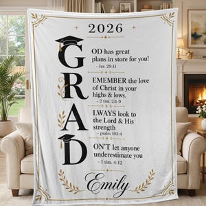 May include: White graduation blanket with the word "GRAD" in large black letters, adorned with graduation caps and floral accents. The blanket includes the year "2026" and inspirational text. The name "Emily" is at the bottom.