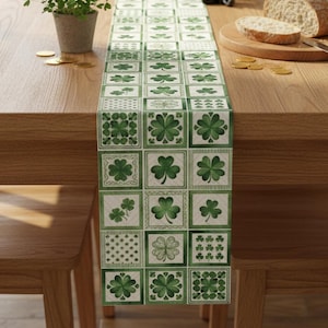 May include: A green and white patterned table runner featuring a repeating design of shamrocks. The runner is rectangular and made of fabric. The shamrocks are in various styles, set against a white and light green background. The table runner is displayed on a wooden table.