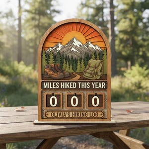 May include: Wooden hiking log with a mountain scene, boots, and backpack. The text "MILES HIKED THIS YEAR" is above three number blocks set to "000". The bottom text reads "OLIVIA'S HIKING LOG". The background shows a forest.