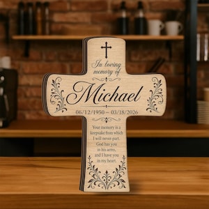 May include: A wooden cross-shaped memorial with the name "Michael" and dates "06/12/1950 - 03/18/2026" engraved. The cross features decorative floral accents and the inscription "In loving memory of". The memorial includes a heartfelt message.